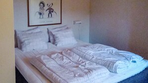 Un lit bien fait, avec une couette blanche et des oreillers, un tableau encadré représentant deux personnages accroché au mur, et un tableau de distribution électrique blanc.
