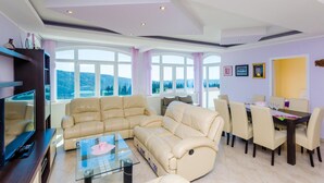 Un salón amplio con un sofá color beige, una mesa de centro de vidrio y una zona de comedor con sillas y una mesa. Amplias ventanas ofrecen vistas al exterior.