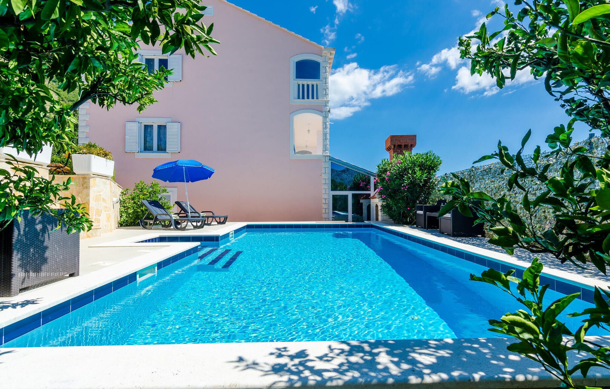 A swimming pool with lounge chairs and an umbrella, surrounded by greenery and a building with a pink facade.