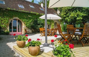 An outdoor patio with a wooden table and chairs, surrounded by potted plants and a pergola with an umbrella.