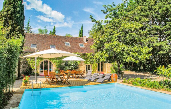 A swimming pool with lounge chairs and umbrellas, surrounded by greenery and a stone building with a tiled roof.