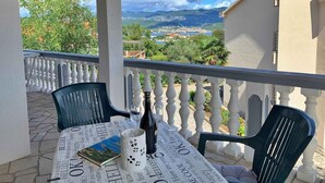 Un balcón con una mesa lista para dos personas, que incluye un libro, una botella y una taza. El balcón da a un paisaje pintoresco con montañas y una masa de agua.