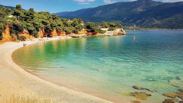 Ein Küstenstrand mit klarem türkisblauem Wasser, einem Sandstrand und einem Hintergrund aus grünen Hügeln und Bergen.