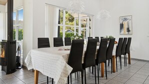 Un coin repas avec une longue table, des chaises noires, une nappe blanche, une cheminée et une fenêtre avec des rideaux.