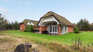 Une maison traditionnelle à toit de chaume, avec une façade en briques, une grande porte vitrée et une pelouse bien entretenue.