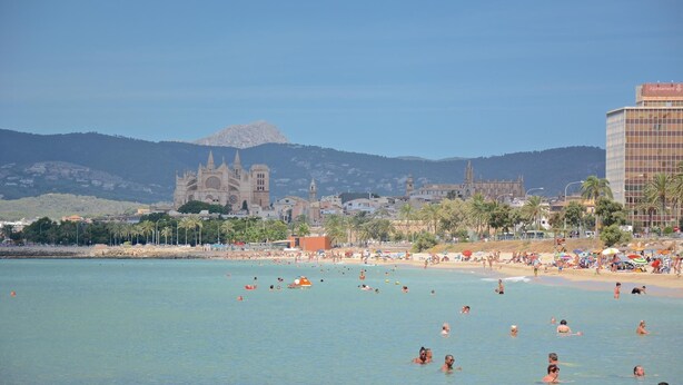 A beach with people swimming and sunbathing, a large cathedral in the background, and a modern building to the right.