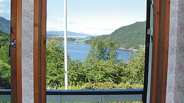 A view through a window with a wooden frame, overlooking a body of water surrounded by greenery and hills. - 3 bedroom amazing home in Etne (Etne)