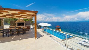 An outdoor patio with a barbecue, poolside chairs, and a view of the sea. - Gorgeous home in Crnja Luka (Crnja Luka)