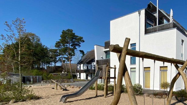 Un'area giochi con scivolo e altalene, circondata da sabbia e alberi, con edifici moderni sullo sfondo.