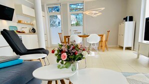 Un soggiorno moderno con un divano, due tavoli rotondi, un tavolino da caffè con fiori, una TV e una cucina sullo sfondo.