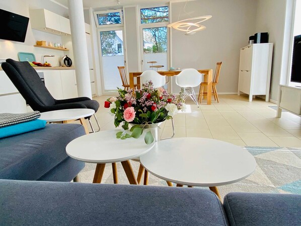 A modern living room with a sofa, two round tables, a coffee table with flowers, a TV, and a kitchen in the background.