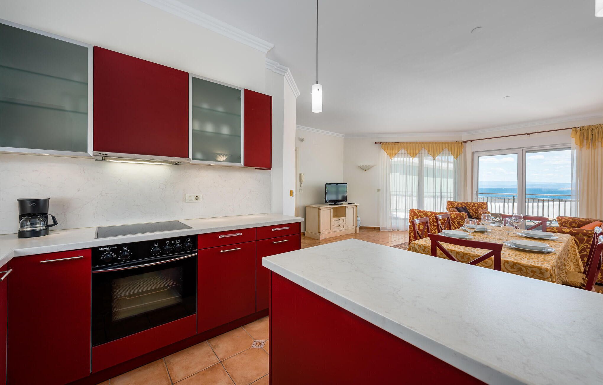 A modern kitchen with red cabinets and a white countertop, a dining area with a table set for four, and a balcony with a view of the sea.