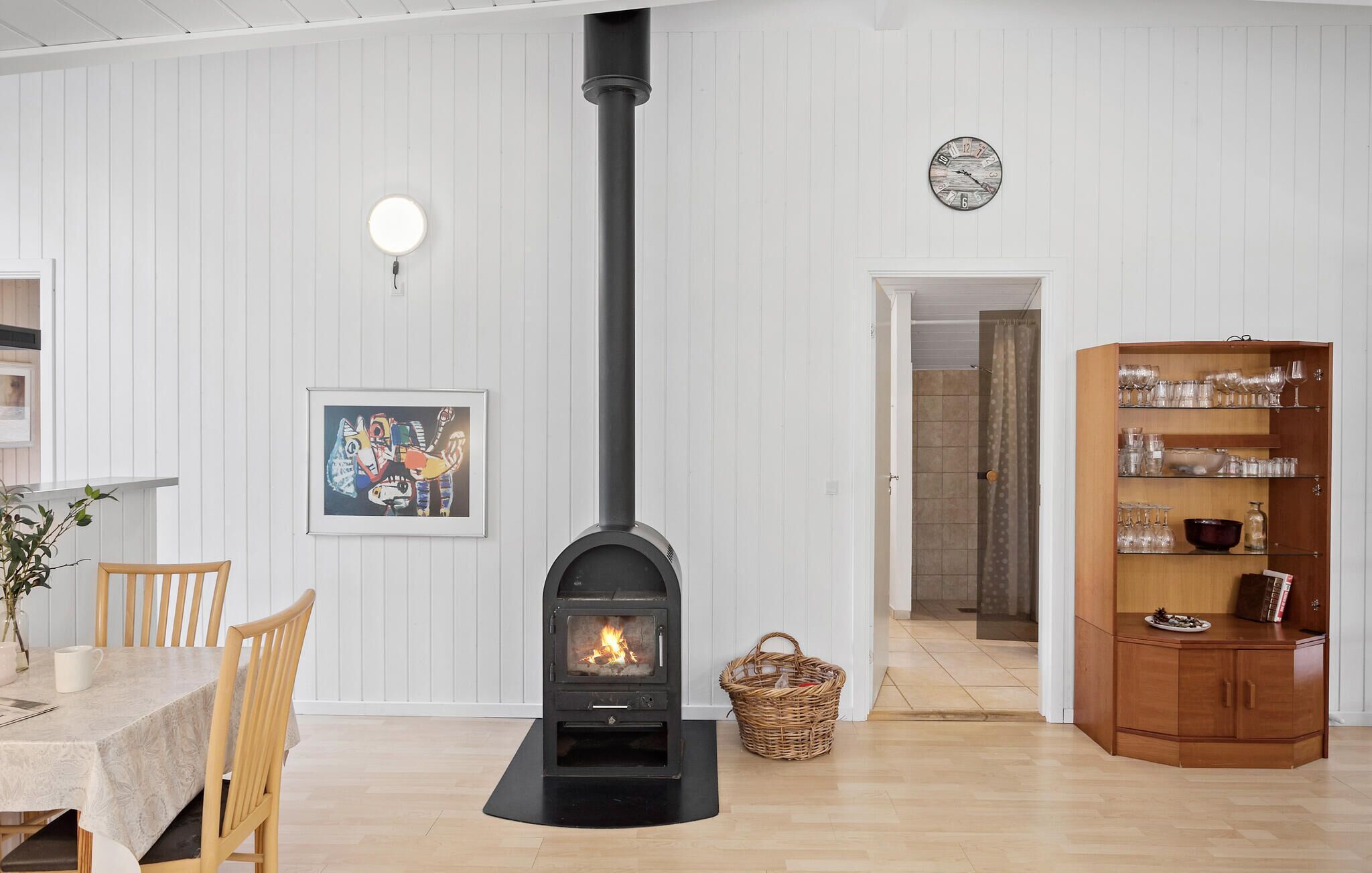Ett vardagsrum med en svart vedkamin, ett matbord med stolar, en inramad abstrakt målning, en korg av rotting och ett träskåp med glasvaror.