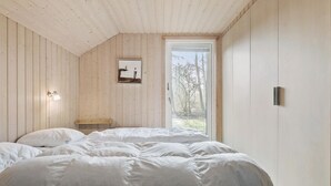 A bedroom with a wooden sloped ceiling, a bed with white bedding, a bedside table, a wall-mounted light, and a window with a view of trees. - Lovely home in Væggerløse with sauna (Væggerløse)