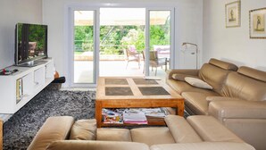 A living room with a sofa, a coffee table with magazines, a television on a white cabinet, and a sliding glass door leading to an outdoor area with chairs and a table.