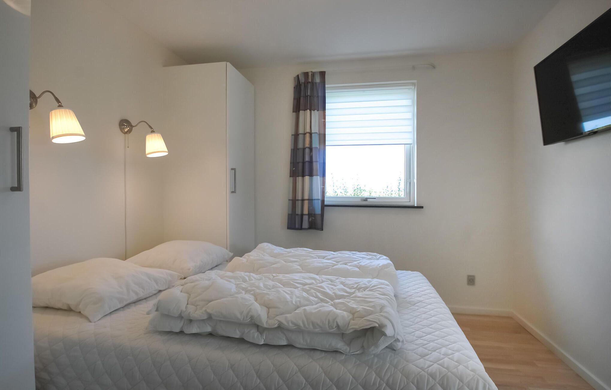 A bedroom with a bed, two wall-mounted lamps, a window with blinds, and a television on the wall.