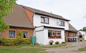 Une maison à deux étages, avec un toit en tuiles, une partie en briques et une façade blanche. On y voit une antenne parabolique, une fenêtre avec un rideau et un petit espace extérieur aménagé avec des chaises et une table.