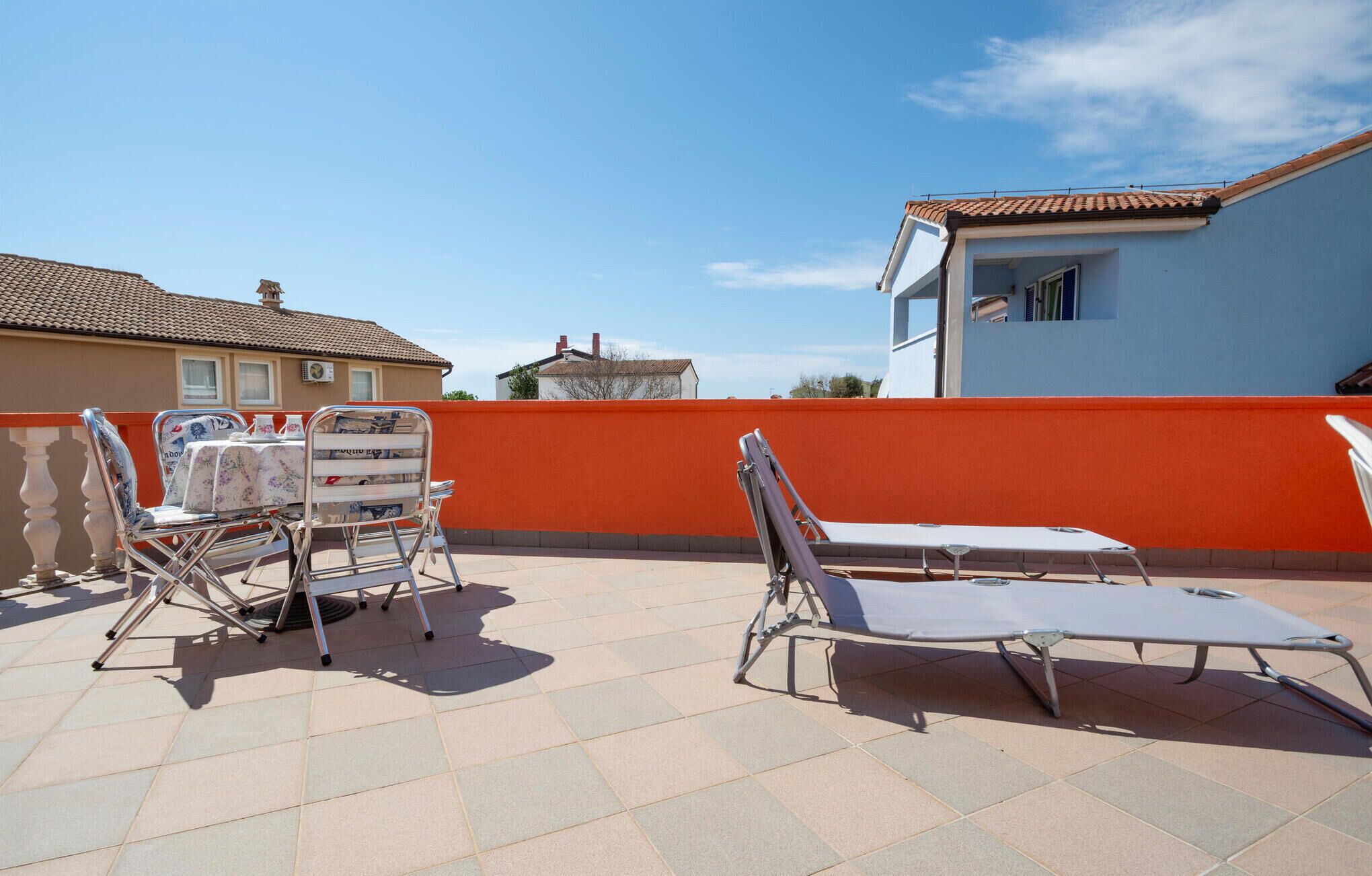 A terrace with two lounge chairs and a table, a red wall, and a blue building in the background.