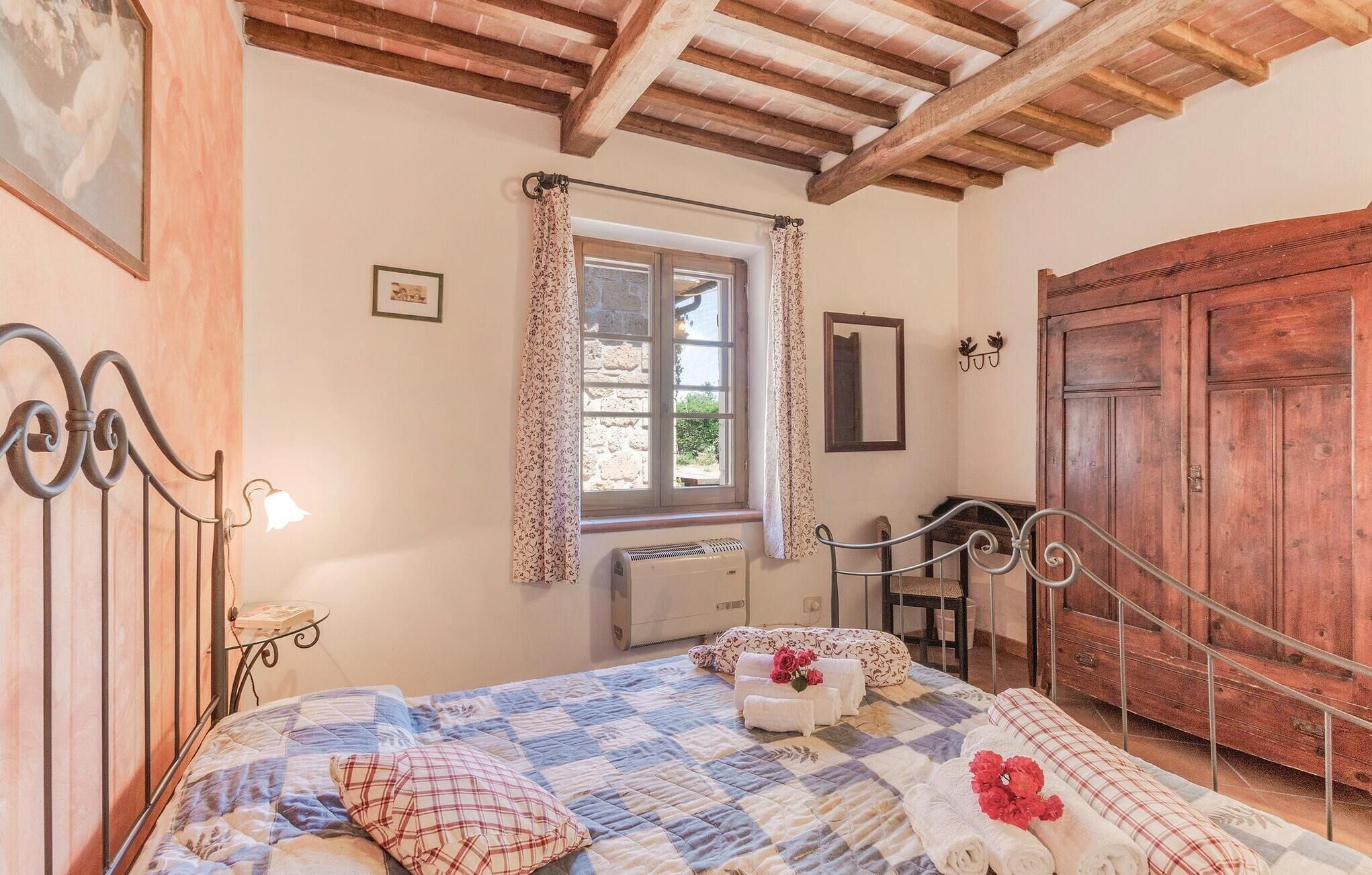 Un dormitorio con cama, ventana con cortinas, radiador, armario de madera y mesita de noche con lámpara.