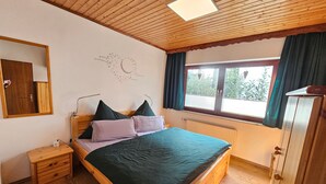 Une chambre avec un plafond en bois, un lit avec une literie verte et violette, une table de chevet, une commode, une fenêtre avec des rideaux et une horloge murale.
