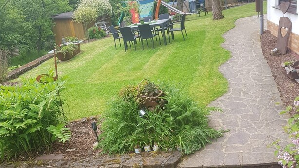 Un chemin de pierres mène à une cour verdoyante agrémentée d’une terrasse en bois et de mobilier d’extérieur.
