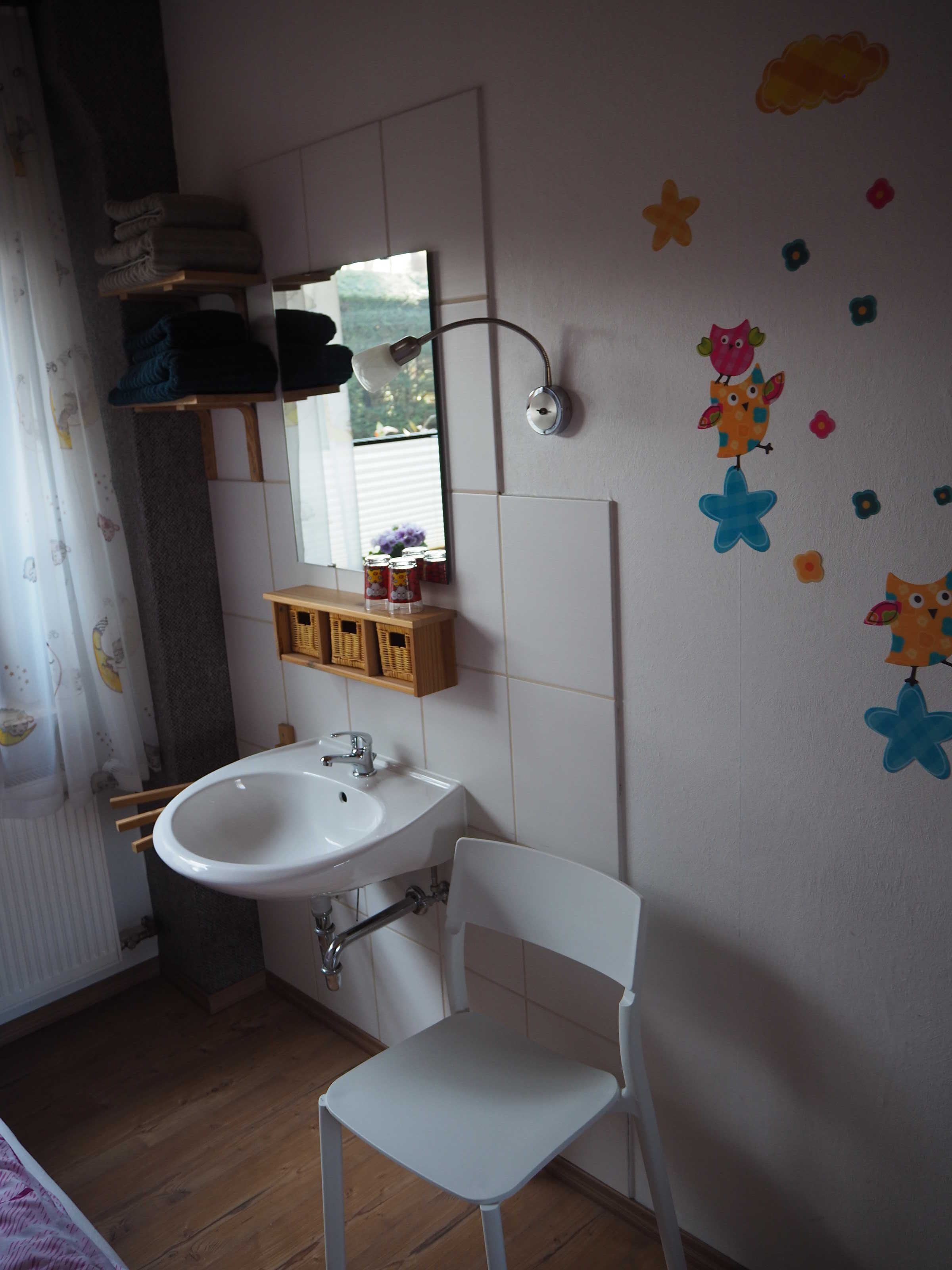 A bathroom with a white sink, a mirror, a white chair, a shelf with towels, and a window.