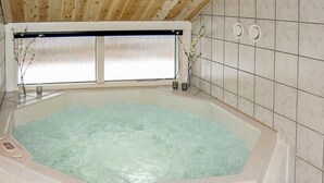 Un bain à remous doté d’un plafond en bois et de murs carrelés, une fenêtre à verre dépoli et un petit vase avec des fleurs posé sur le rebord de la fenêtre.