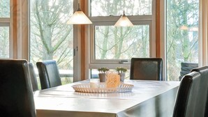 Un coin repas avec une table dressée pour six personnes, de grandes fenêtres donnant sur la forêt et deux lampes suspendues.