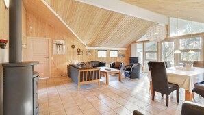 Une pièce chaleureuse et bien éclairée, avec un plafond haut, des murs en bois, une cheminée et un coin repas comprenant une table et des chaises.