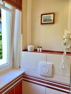 Une salle de bain avec une fenêtre, un tableau encadré au mur, un téléphone et des articles de toilette rangés sur une étagère.