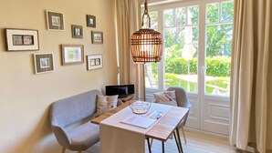 A room with a sofa, a table with a laptop, framed pictures on the wall, and a view of greenery outside. - Apartment 1 - Villa Amelie (Binz)