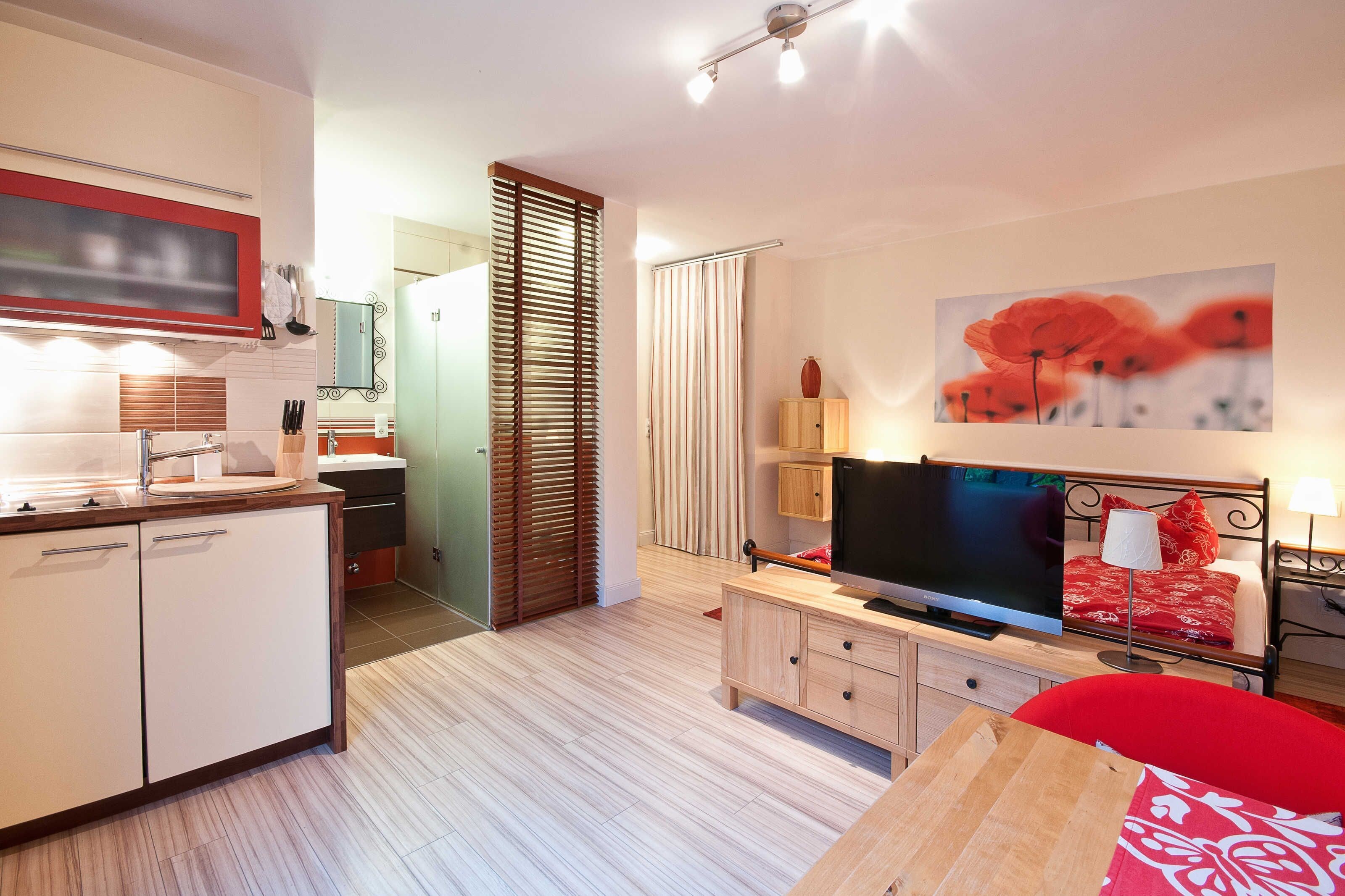 Eine moderne Küche mit Fernseher, ein Essbereich mit einem roten Sessel und ein Schlafzimmer mit Fernseher und einer roten Bettdecke.