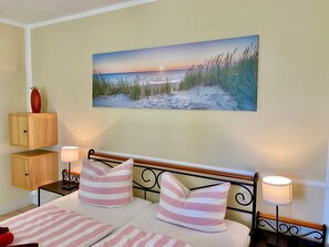 Ein Schlafzimmer mit einem Bett, Kissen, Nachttischen, einer Lampe, einem Bild von einem Strand und einem Holzschrank.