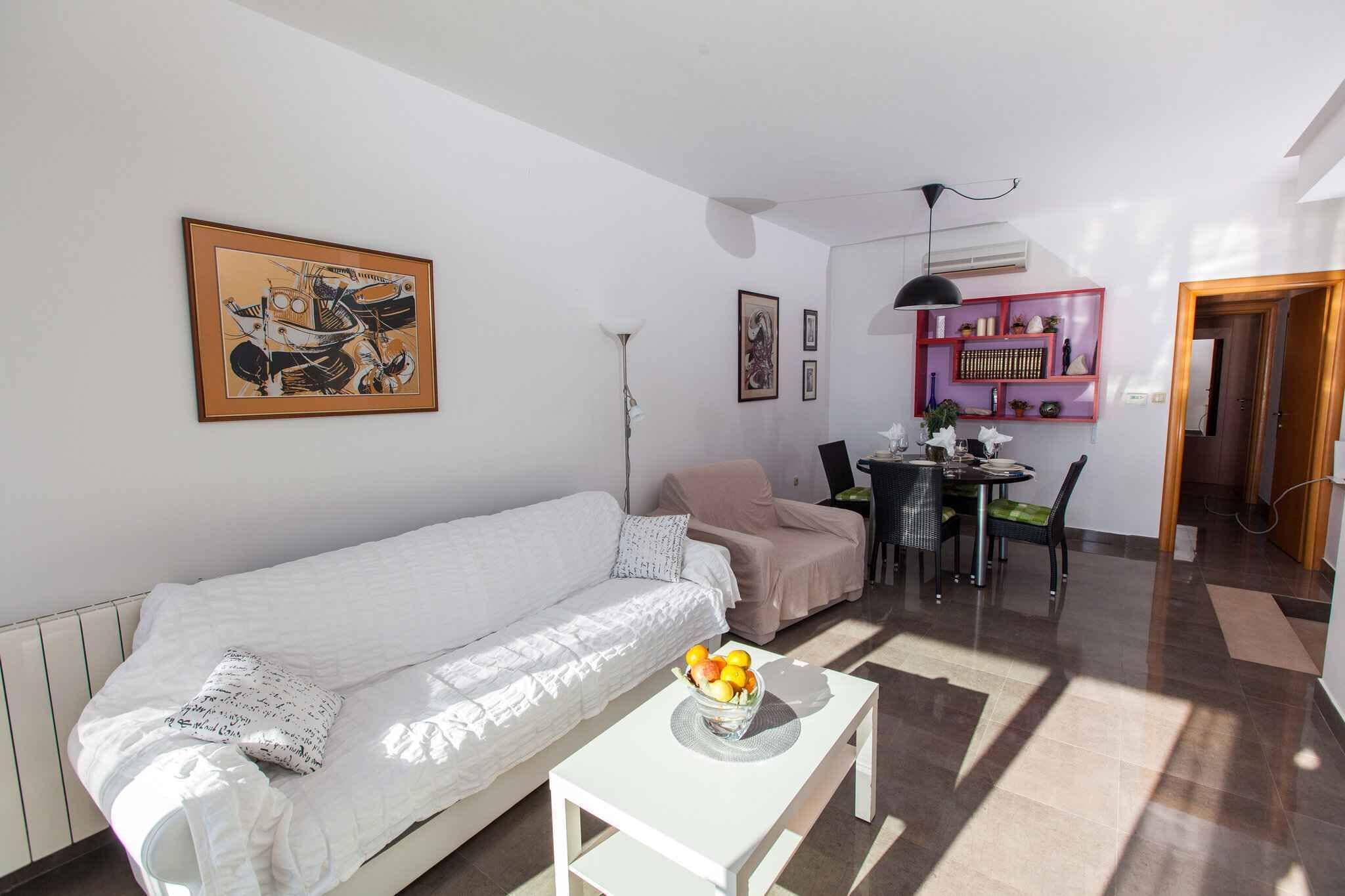 Un salon moderne avec un canapé blanc, une table à manger avec des chaises et une table basse sur laquelle sont disposés des fruits. Un tableau orne le mur, et une bibliothèque contient des livres et des objets décoratifs.