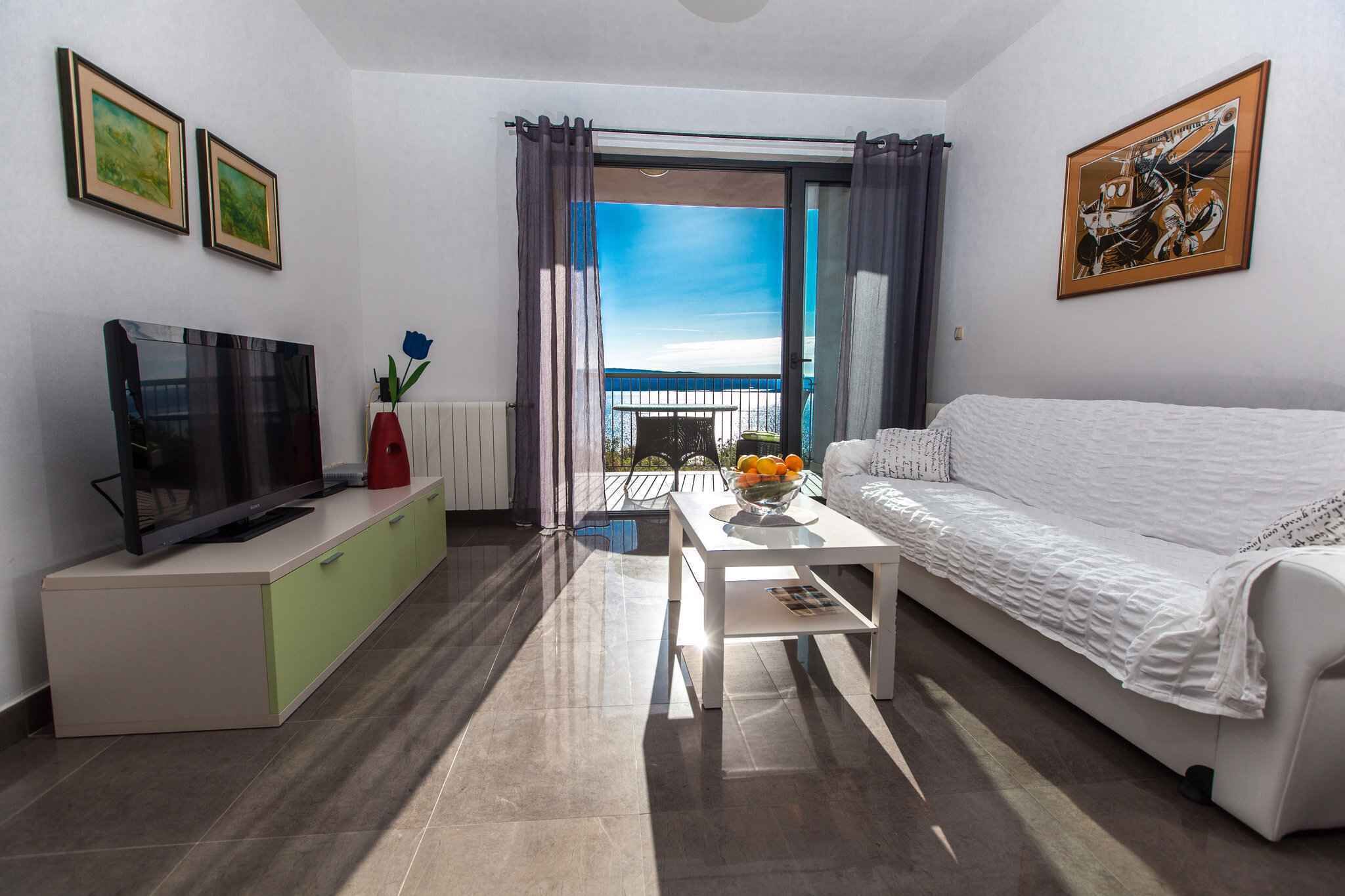 A modern living room with a flat-screen TV, a white sofa, a coffee table with fruit, and a balcony with a view.