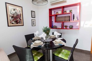 A dining area with a round table set for six, surrounded by wicker chairs with green cushions. A red shelf with decorative items and books is mounted on the wall. - Holiday apartment mit Meerblick (Dramalj)