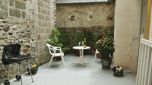 Un petit espace extérieur aménagé en patio, avec un barbecue, une chaise en plastique blanche, une petite table ronde sur laquelle se trouvent une bouteille et un verre, ainsi que des plantes en pot.