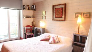 A bedroom with a large bed, a desk with a chair, a bookshelf, and a window with blinds.