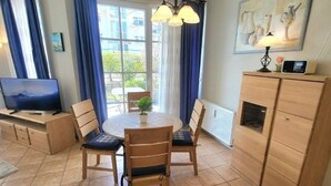 A dining area with a round table, four chairs, a television, a cabinet, and a window with curtains. - (49) 3-room apartment with south-facing terrace - Appartementhaus Atlantik (Kühlungsborn)