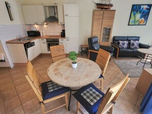 A compact kitchen with white cabinets, a microwave, and a stove. There is a dining area with a round table and chairs, and a sofa in the living room. - (49) 3-room apartment with south-facing terrace - Appartementhaus Atlantik (Kühlungsborn)