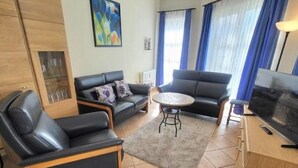 A living room with two black leather sofas, a round coffee table, a TV on a wooden cabinet, and a glass cabinet with glasses. - (49) 3-room apartment with south-facing terrace - Appartementhaus Atlantik (Kühlungsborn)