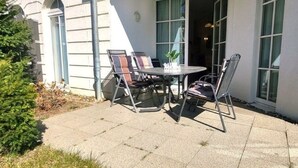 An outdoor patio with a table and chairs, a potted plant, and a building with large windows and a door. - (49) 3-room apartment with south-facing terrace - Appartementhaus Atlantik (Kühlungsborn)