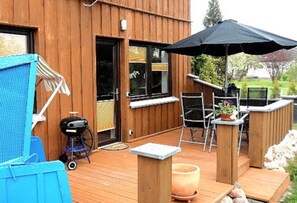 Une terrasse en bois avec un barbecue, des sièges d’extérieur et un grand parasol.