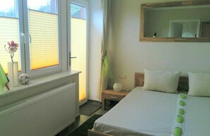 Ein Schlafzimmer mit Bett, einem Fenster mit Jalousien, einem Heizkörper, einem kleinen Tisch mit Blumenvase und einem Spiegel an der Wand.