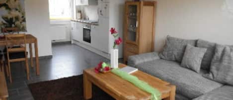 Una sala de estar con un sofá gris, una mesa de centro de madera, una zona de comedor con una mesa y sillas, y una cocina al fondo.
