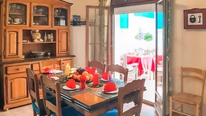 Un comedor con una mesa de madera dispuesta para cuatro personas, rodeada de sillas. Un aparador con puertas de cristal está a la izquierda, y una puerta a la derecha da al exterior.