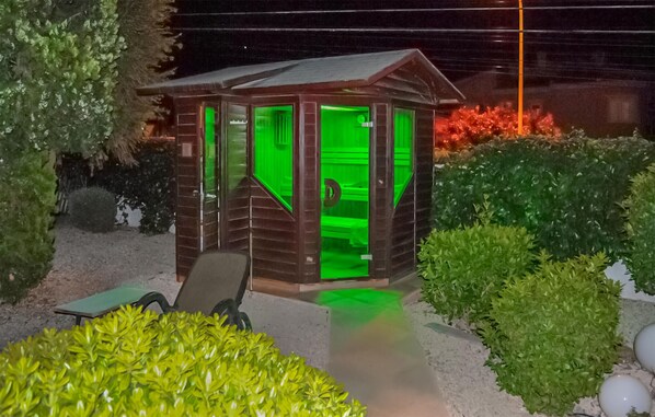 A small wooden cabin with a green-lit interior, surrounded by well-maintained bushes and a gravel path. - Awesome home in Rojales with sauna (Rojales)