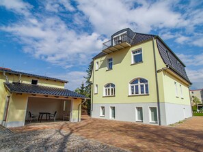 Ein zweistöckiges Gebäude mit einem Balkon im oberen Geschoss, einer überdachten Terrasse mit Tisch und Stühlen sowie einer gepflasterten Auffahrt.