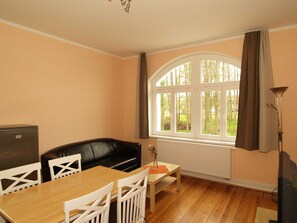 Una habitación con una mesa de comedor de madera, sillas blancas, un sofá de cuero negro, un radiador, una ventana con cortinas y una lámpara de pie.