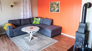 A living room with a grey sectional sofa, a small round table, a red and white patterned rug, a black wood-burning stove, and a wall with an orange panel and a framed picture. - Lovely home in Sveio with WiFi (Sveio)